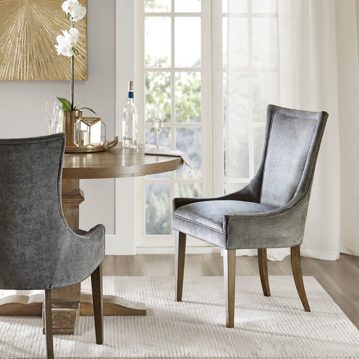 A modern dining area features a round wooden table, an Ultra - Elegant Dining Side Chair (Set of 2) upholstered in gray, a white rug, and a gold decorative centerpiece. Sunlight streams through large glass doors, brightening the space.