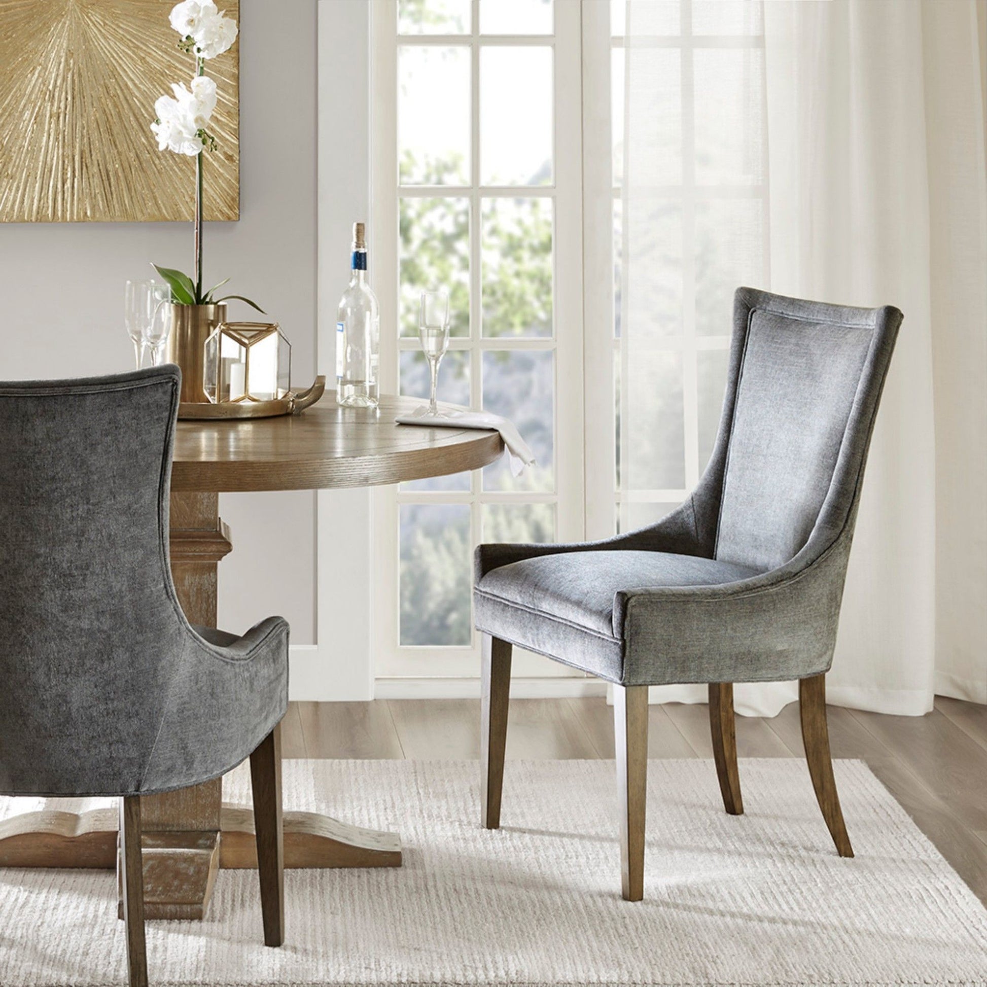A modern dining area features a round wooden table, an Ultra - Elegant Dining Side Chair (Set of 2) upholstered in gray, a white rug, and a gold decorative centerpiece. Sunlight streams through large glass doors, brightening the space.