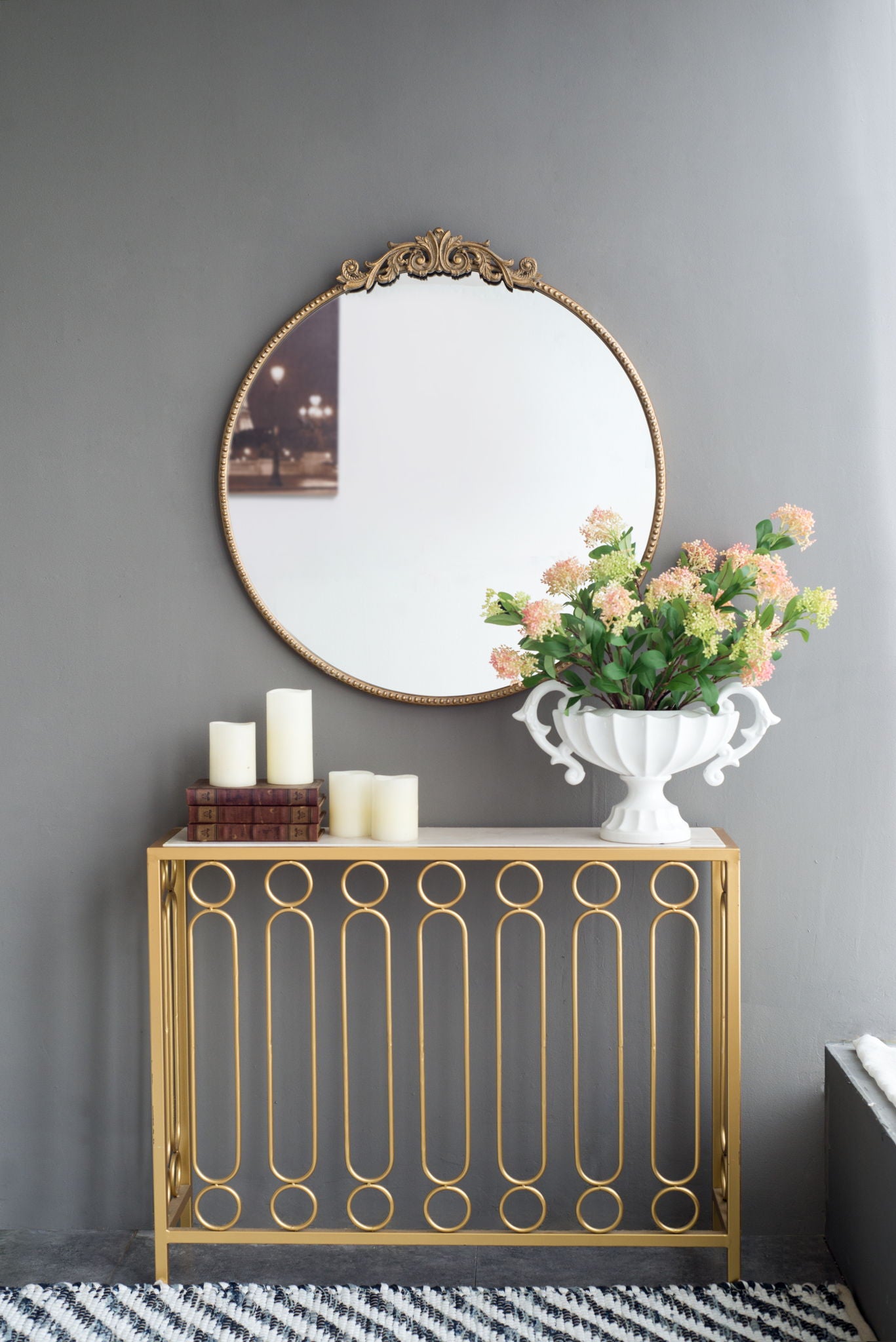A Classic Design Mirror With Baroque Inspired Frame hangs above a gold table with stacked books, pillar candles, and a white vase of pink and green flowers. The gray wall and striped rug complete the elegant entryway scene.