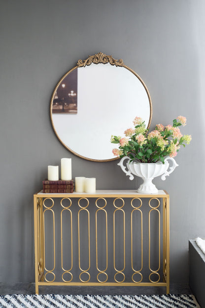 A Classic Design Mirror With Baroque Inspired Frame hangs above a gold table with stacked books, pillar candles, and a white vase of pink and green flowers. The gray wall and striped rug complete the elegant entryway scene.
