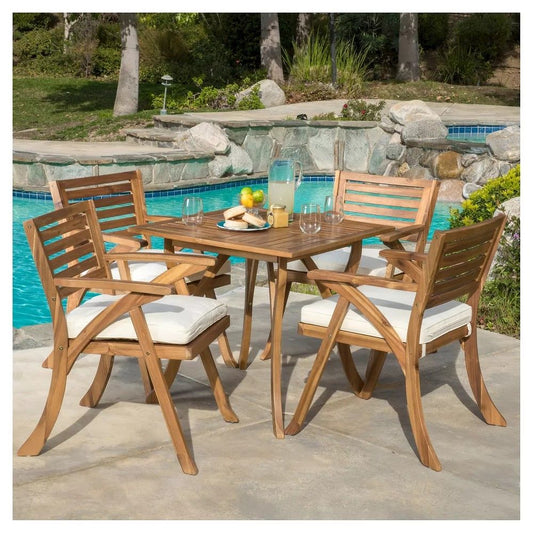 The Outdoor Hermosa 5 Piece Wood Dining Set With Cushions in teak and beige is arranged beside a pool, featuring a table with lemonade, glasses, fruit, and a bowl. Greenery and rocks provide a scenic backdrop.