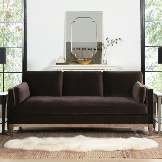 A modern living room features the Knox - Modern Farmhouse Sofa in dark brown velvet, a white fur rug, and side tables. Behind the sofa sits a white mantel with decor, a mirror, and green plants. Large windows border the space.