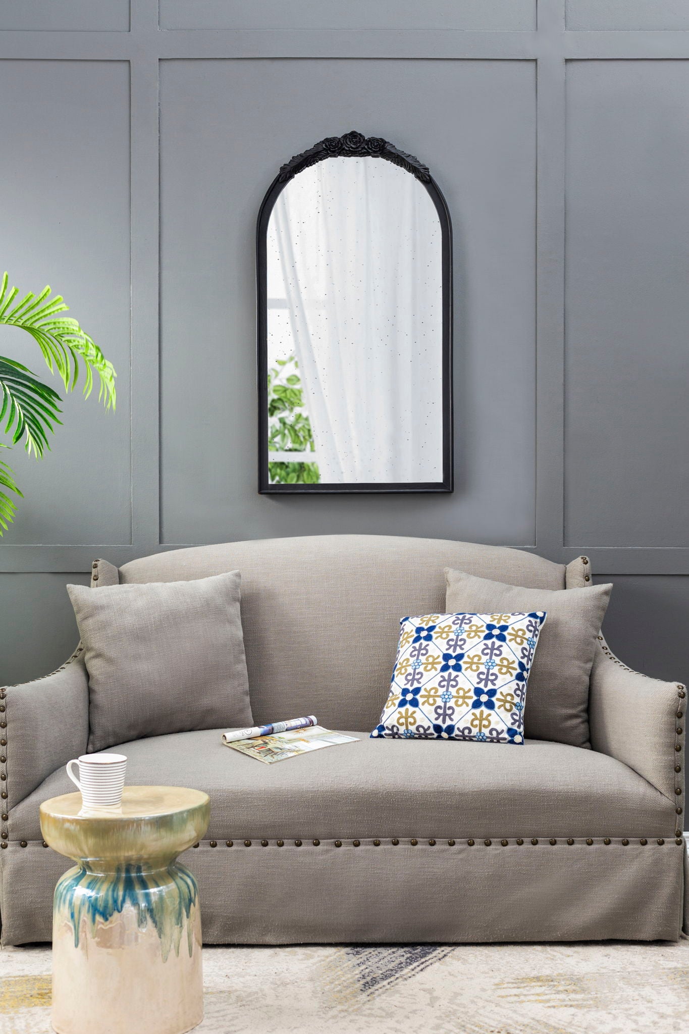 A gray sofa with two cushions sits against a gray paneled wall, featuring the Hand Carved Rose Antique Mirror Frame, Wood Arch Mirror Wall Decor For Living Room above. A small round side table holds a white mug and magazine, with a plant to the left.