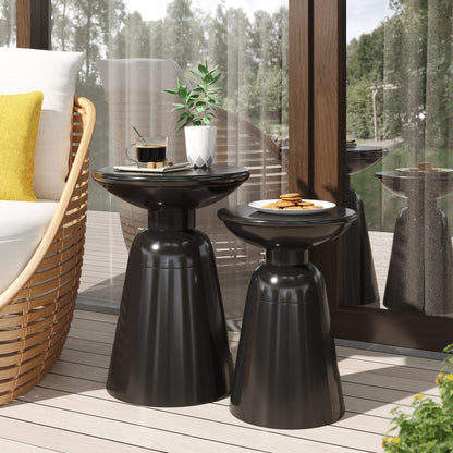 Two Serenita Outdoor Side Tables with flared design are on a patio—one holds coffee and a plant, the other has cookies—set beside a wicker chair with a yellow pillow and a large window reflecting the scene.