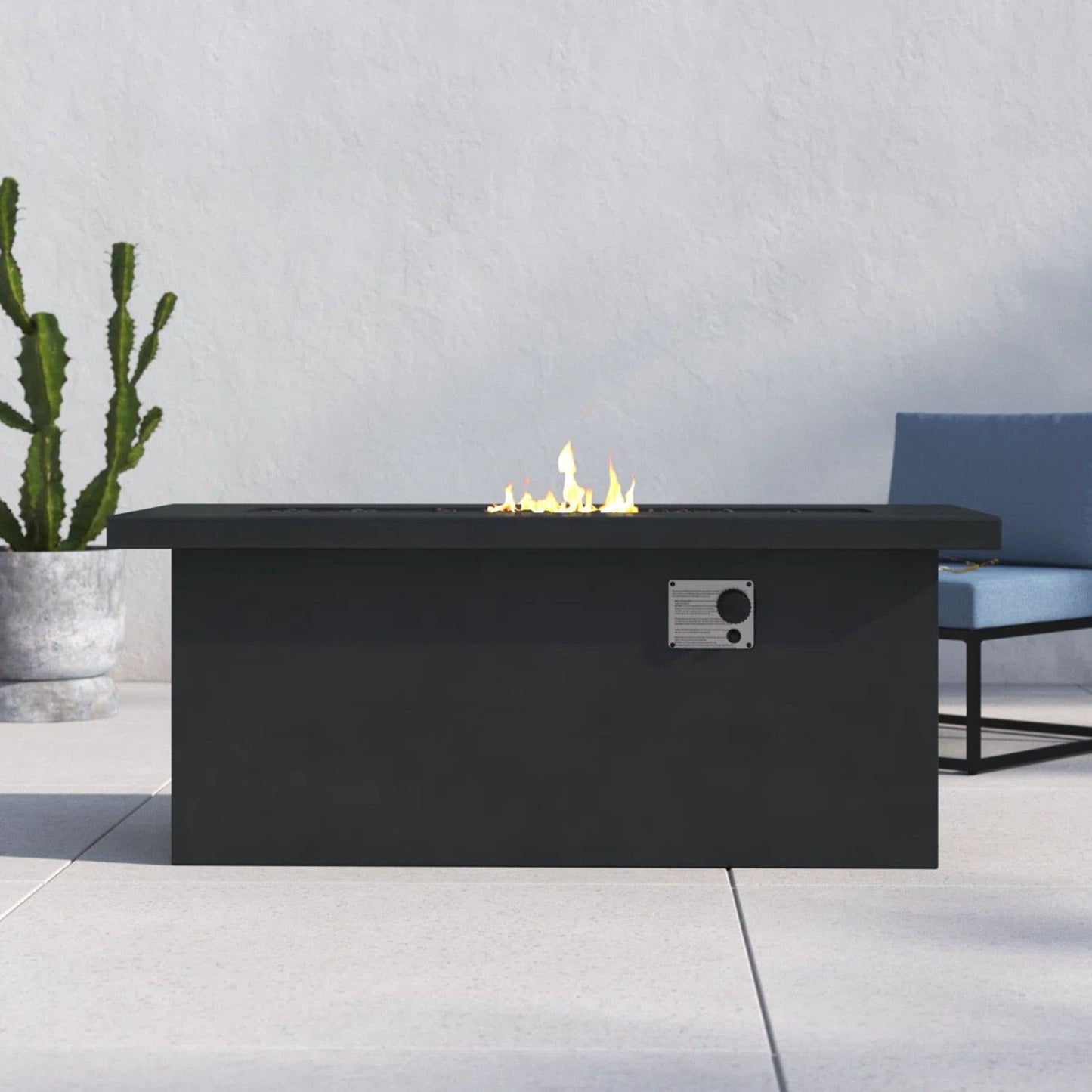 A Propane / Gas Outdoor Fire Pit Table with visible flames stands on a patio. A potted cactus sits to the left, while a blue-cushioned chair is on the right, all set against a light gray wall.