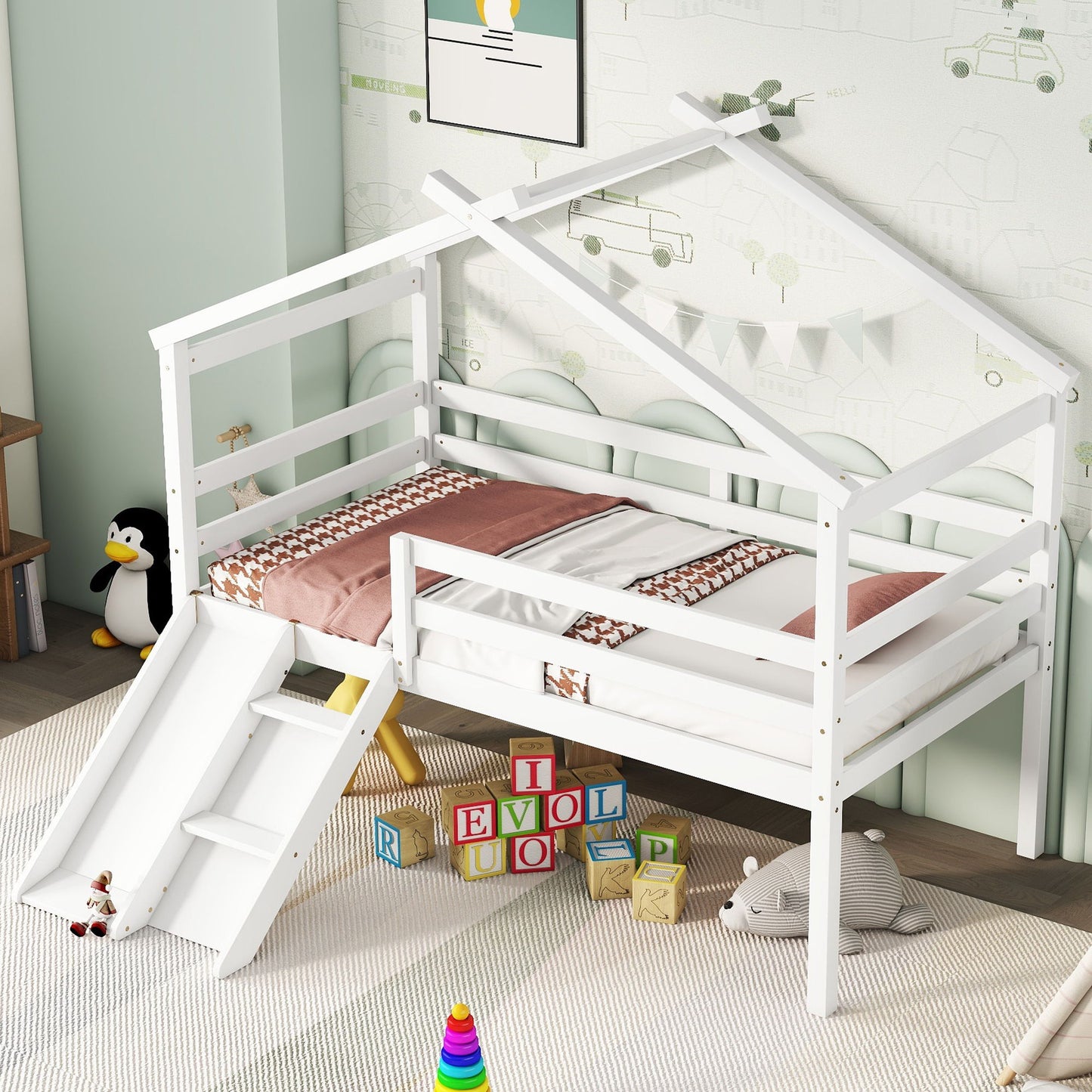 A Low Loft House Bed With Slide, Ladder, Safety Guardrails, and a House Roof Frame sits in a playful bedroom. Alphabet blocks, toys, a plush bear, and penguin decor are scattered on a light rug beside the bed.