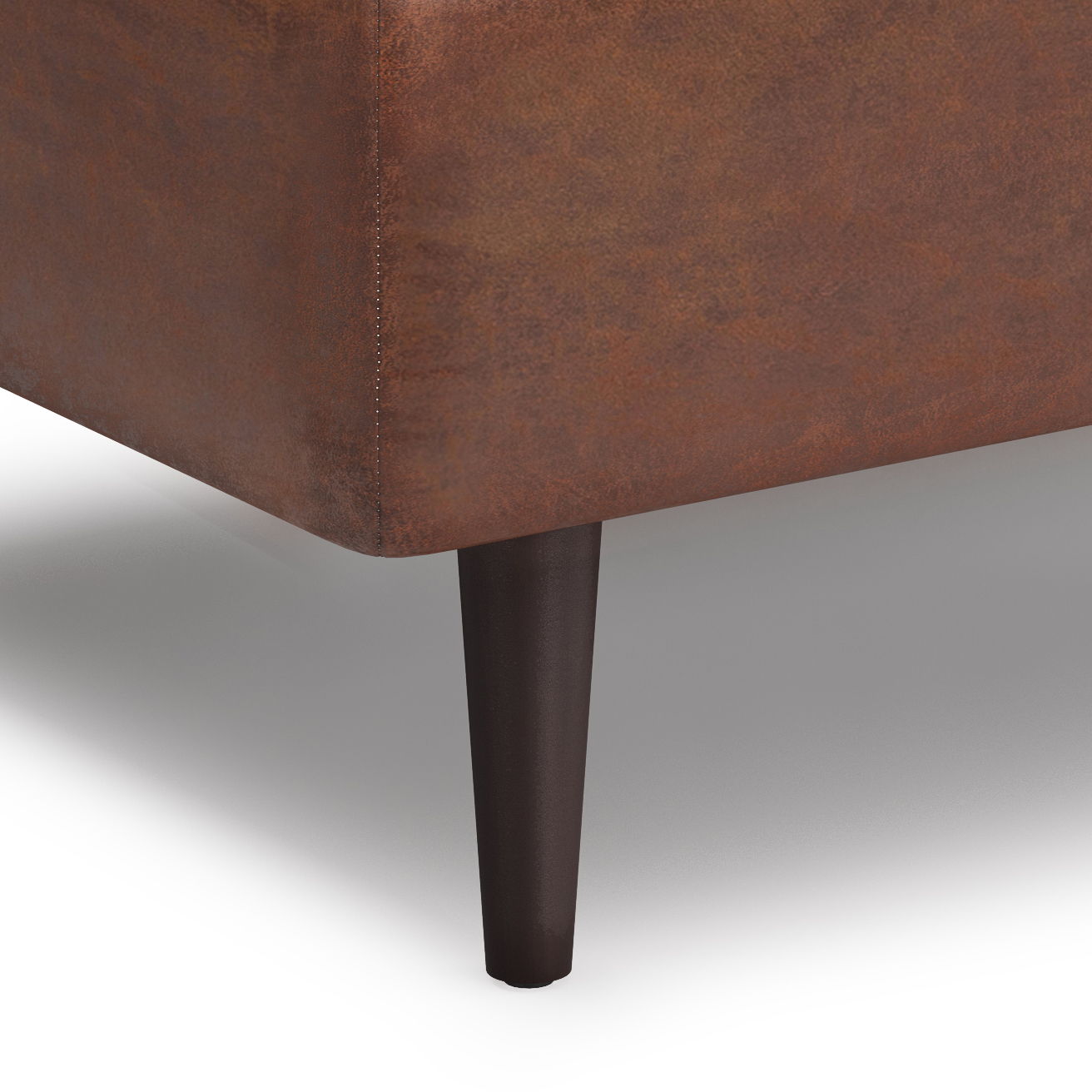 Close-up of the corner of the Shay - Large Square Coffee Table Storage Ottoman Mid-Century Style, featuring brown leather with visible stitching and one dark wooden tapered leg on a white background.