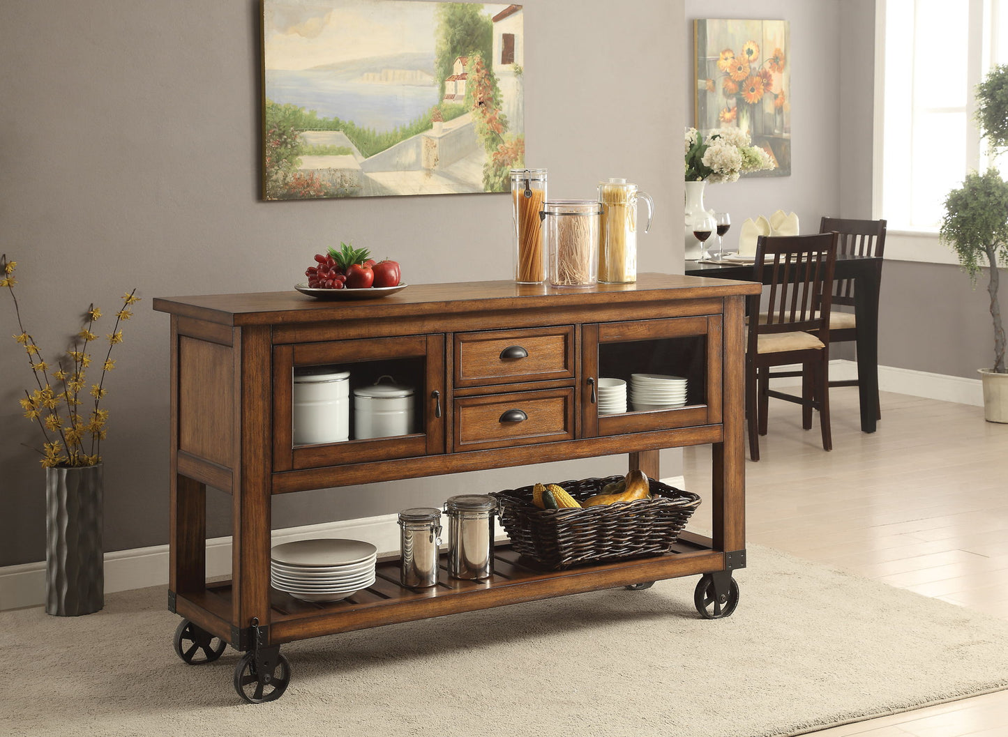 The Kadri Kitchen Cart in Distressed Chestnut, with drawers and shelves for jars, plates, fruit baskets, and glass canisters, stands on wheels in a bright dining area featuring wall art, flowers, and a dining table.