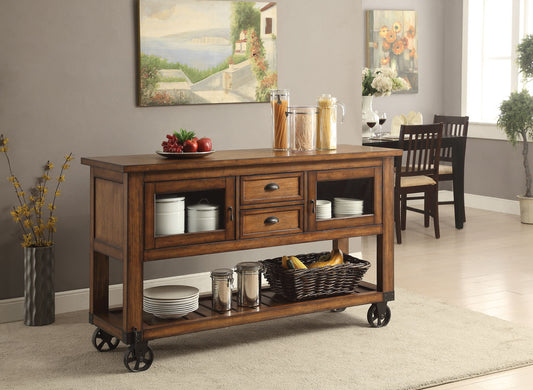The Kadri Kitchen Cart in Distressed Chestnut, with drawers and shelves for jars, plates, fruit baskets, and glass canisters, stands on wheels in a bright dining area featuring wall art, flowers, and a dining table.