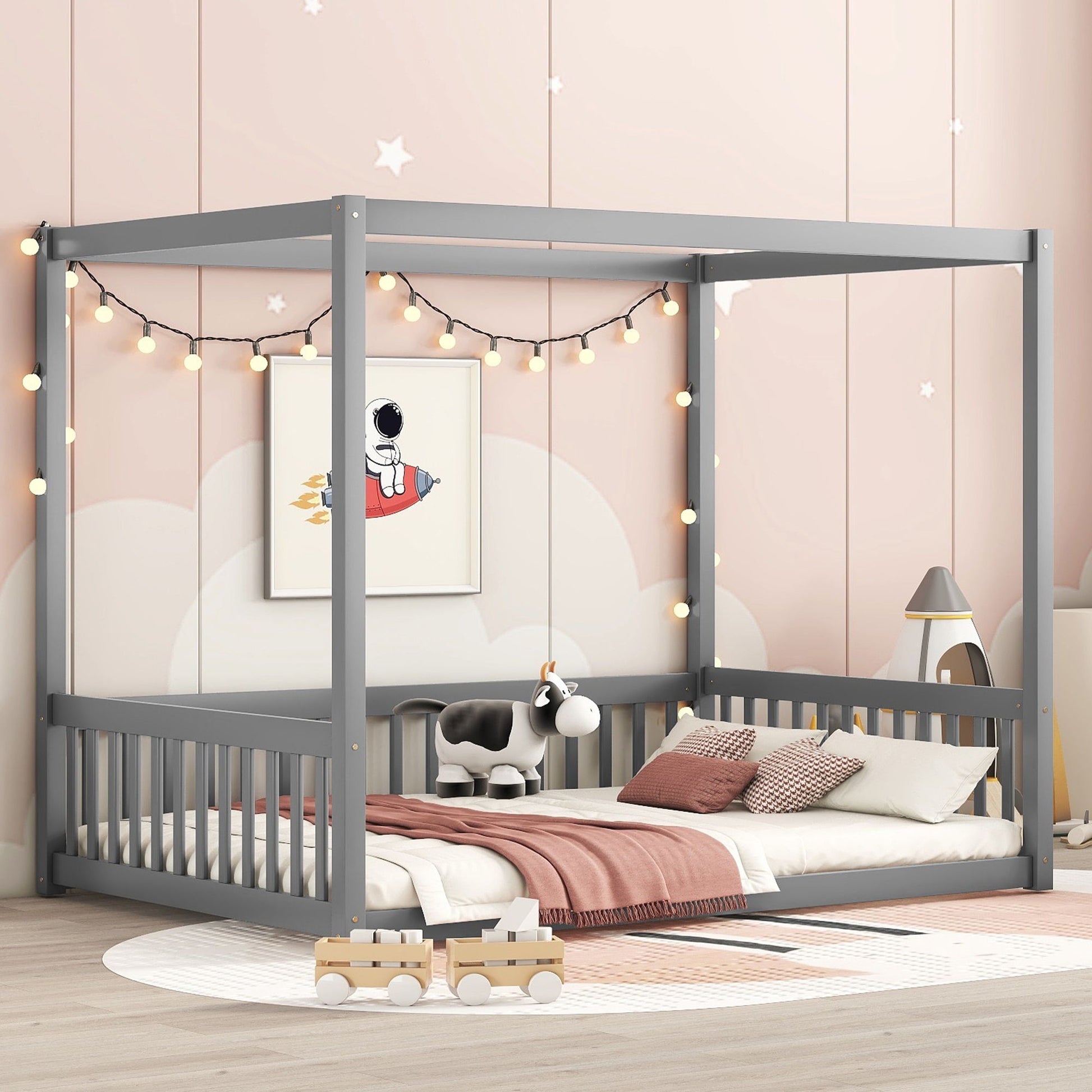 A cozy children’s bedroom features the Canopy Frame Floor Bed With Fence, Guardrails in gray with string lights, pillows, a plush cow toy, wooden trucks, pastel decor, pink walls with a rocket print, and white cloud accents.