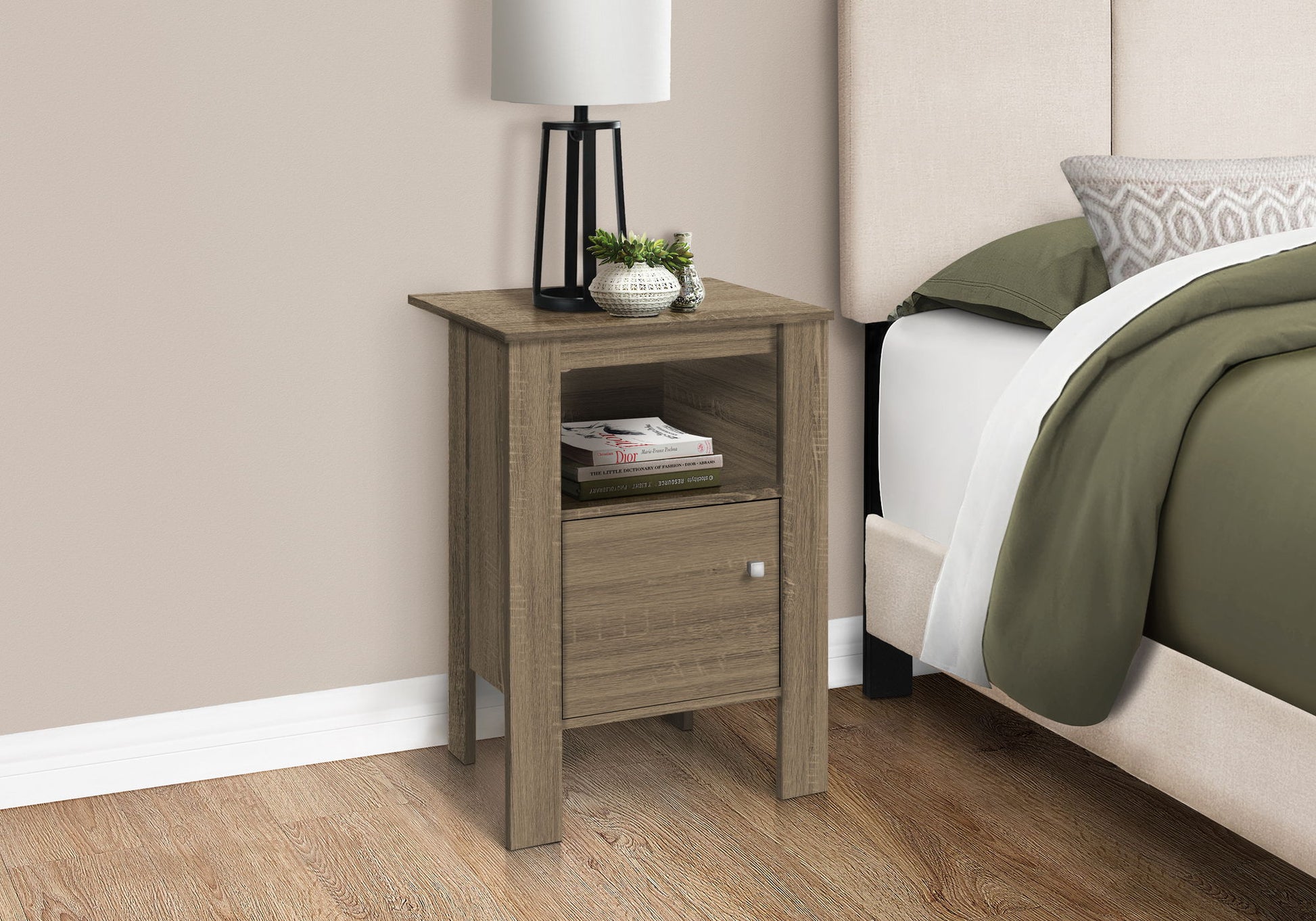 An Accent Nightstand with storage and a transitional design, featuring a drawer and open shelf holding a lamp, plant, and book, stands beside a bed with green and white bedding in a light-walled room with wood flooring.