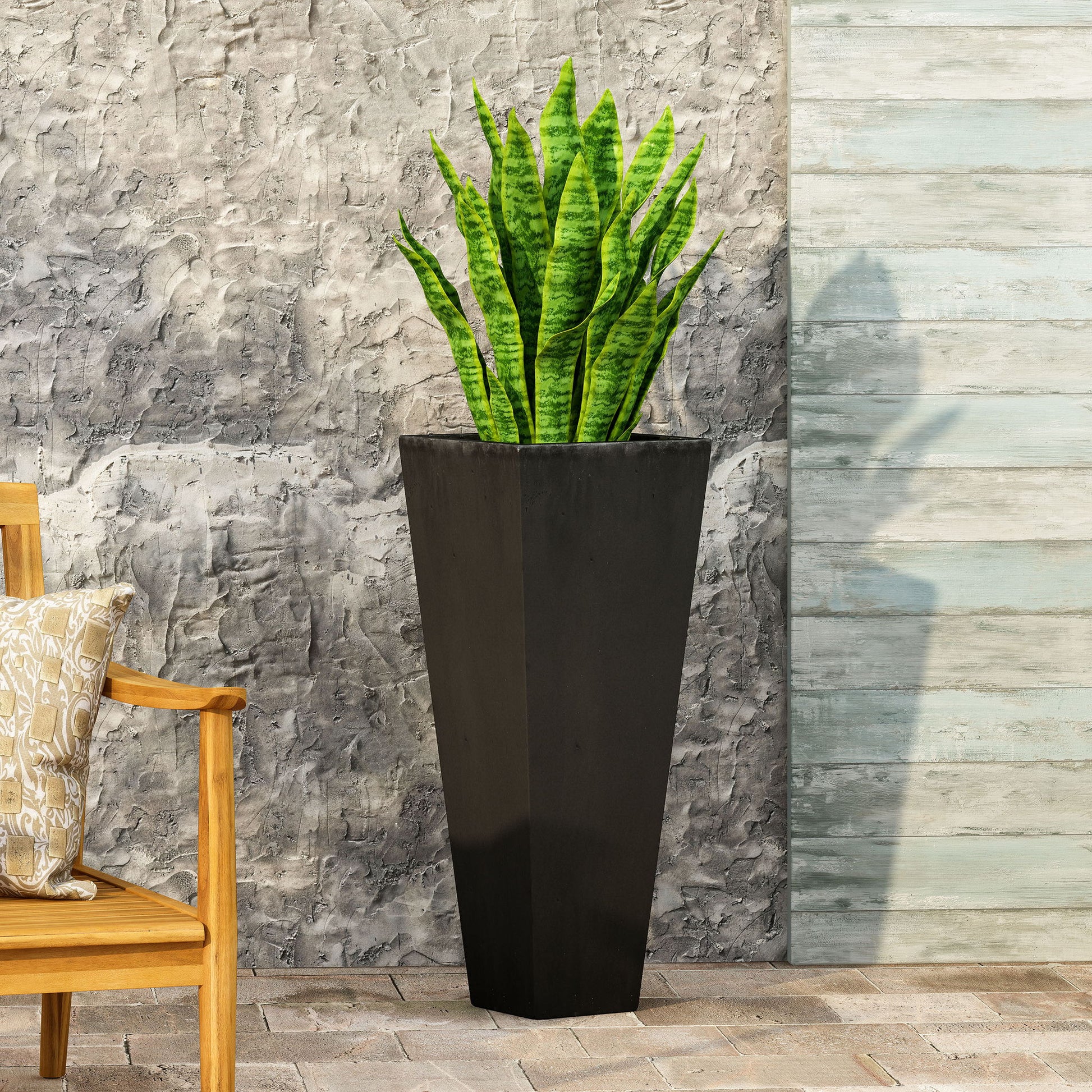 An Outdoor Modern Cast Stone Planter with a snake plant stands on outdoor tiles by a textured concrete wall, next to a wooden chair with a patterned cushion.