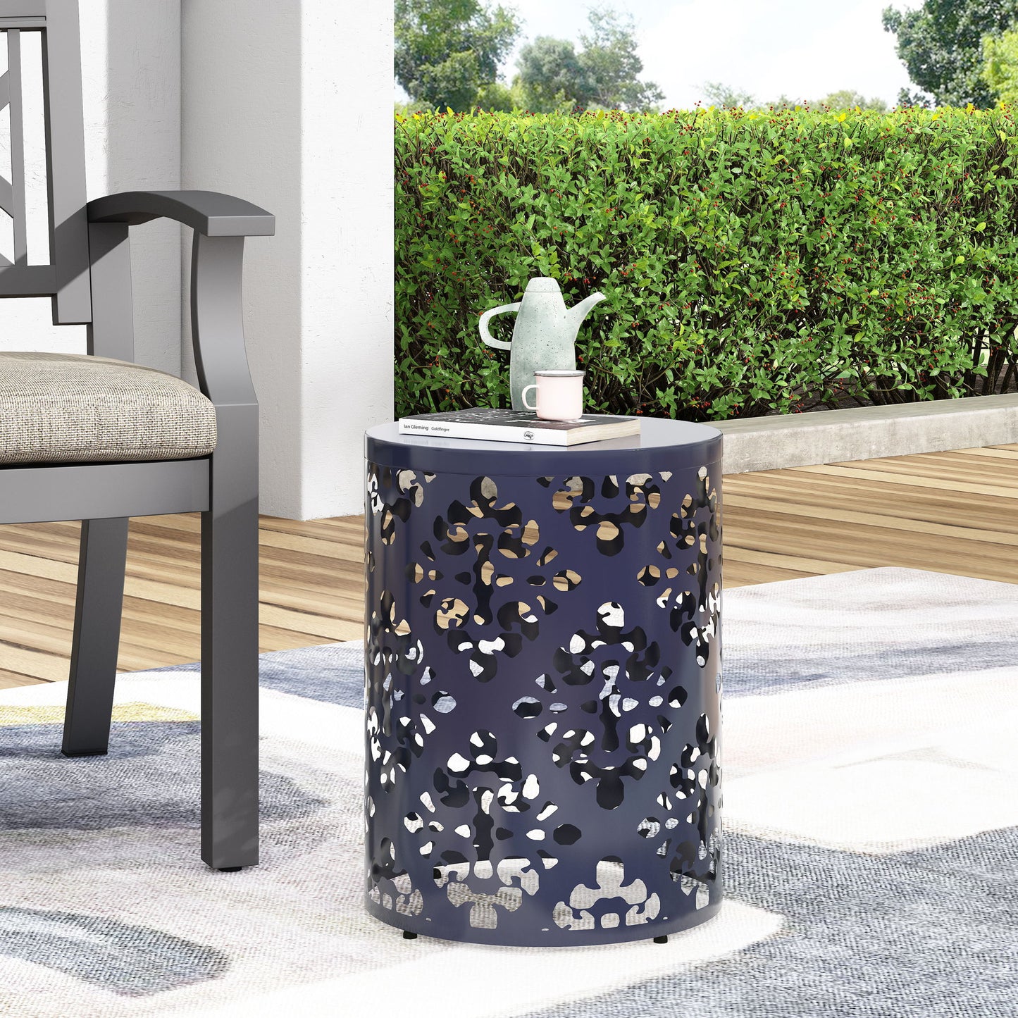 The Outdoor Metal Modern Design End Table, small and round with decorative cutouts, sits on a patio rug next to a cushioned outdoor chair. A teapot, cup, and book rest on top, with green bushes and trees visible in the background.