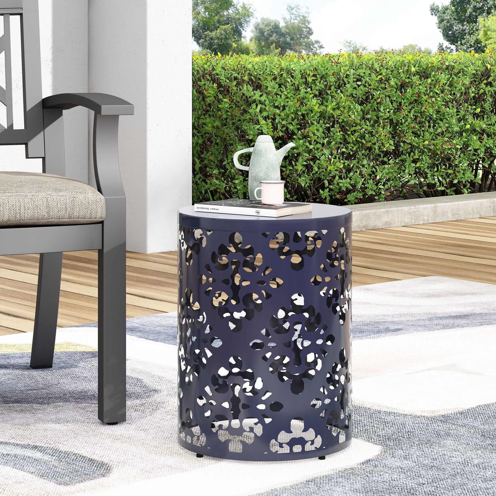 The Outdoor Metal Modern Design End Table, small and round with decorative cutouts, sits on a patio rug next to a cushioned outdoor chair. A teapot, cup, and book rest on top, with green bushes and trees visible in the background.