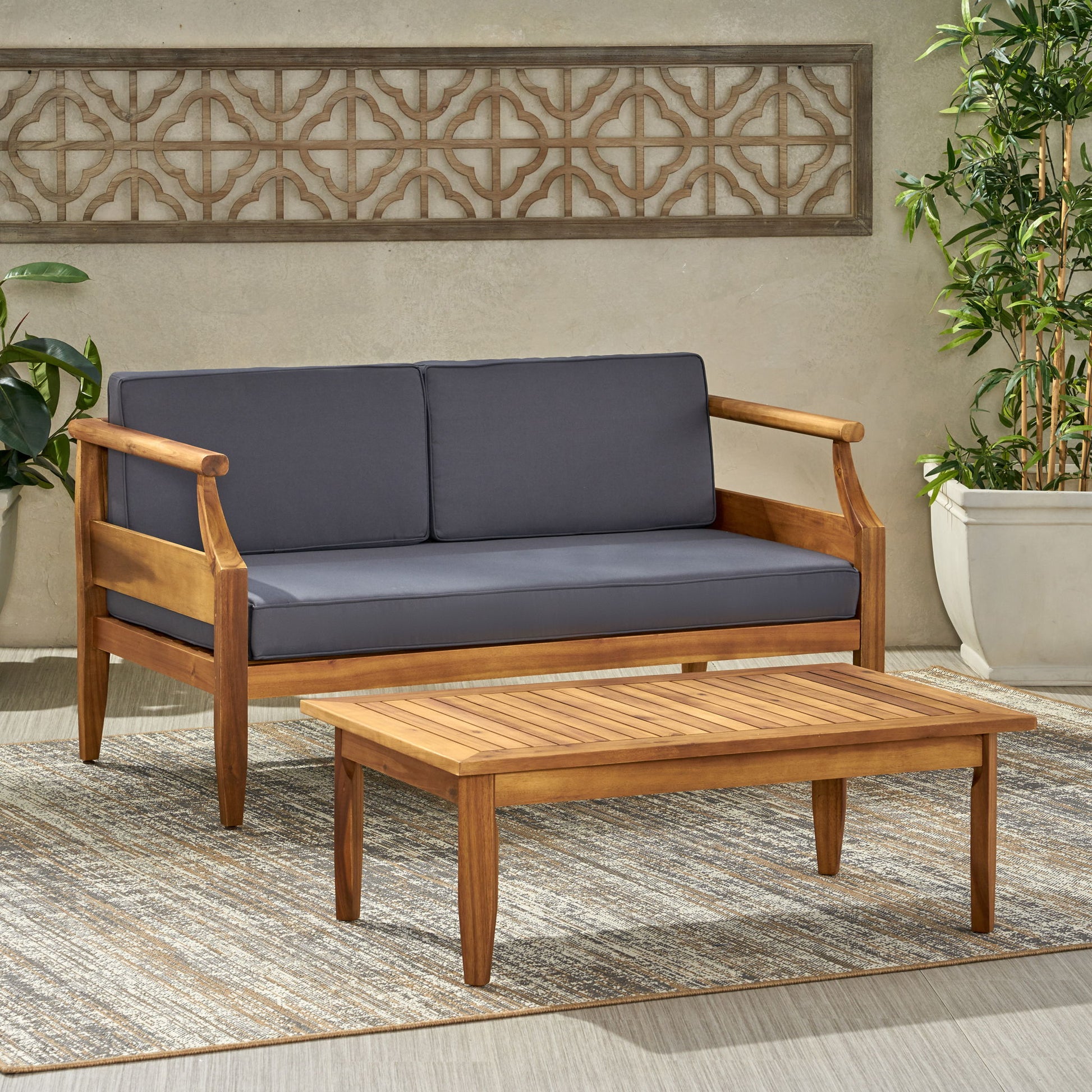 The Aston - 2 Piece Wood Outdoor Patio Conversation Seating Set With Cushions features a wooden loveseat with dark gray cushions and a matching coffee table on a striped rug, accented by potted plants and a decorative wall panel.