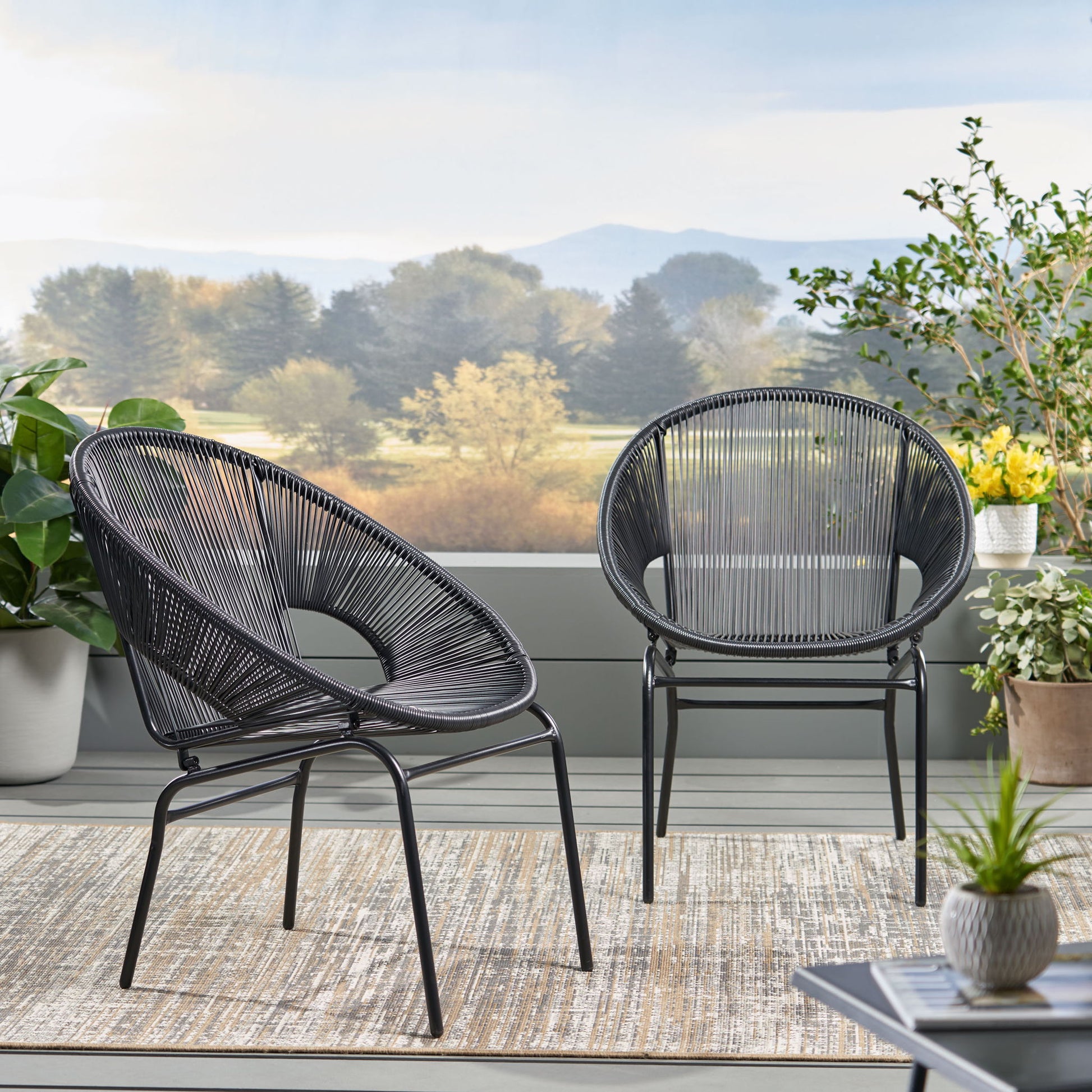 Two Nusa - Modern Chair Outdoor Seating pieces with circular woven designs sit on a patio rug, surrounded by potted plants. Trees and mountains create a scenic backdrop.
