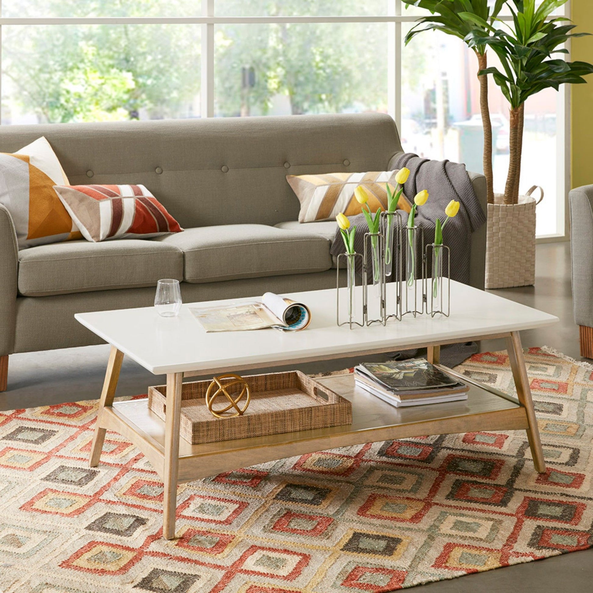 A modern living room features a gray sofa, colorful geometric rug, and a Modern Table With Extra Storage holding a glass, magazines, baskets, and yellow tulips in metal vases, all set before large windows.