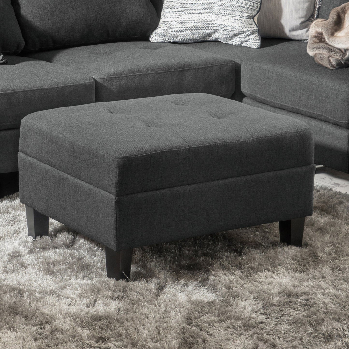 A Classic Ottoman With Birch Wood Legs And Fabric Upholstery in dark gray sits on a light gray shaggy rug in front of a matching sectional sofa with cushions and a throw blanket.
