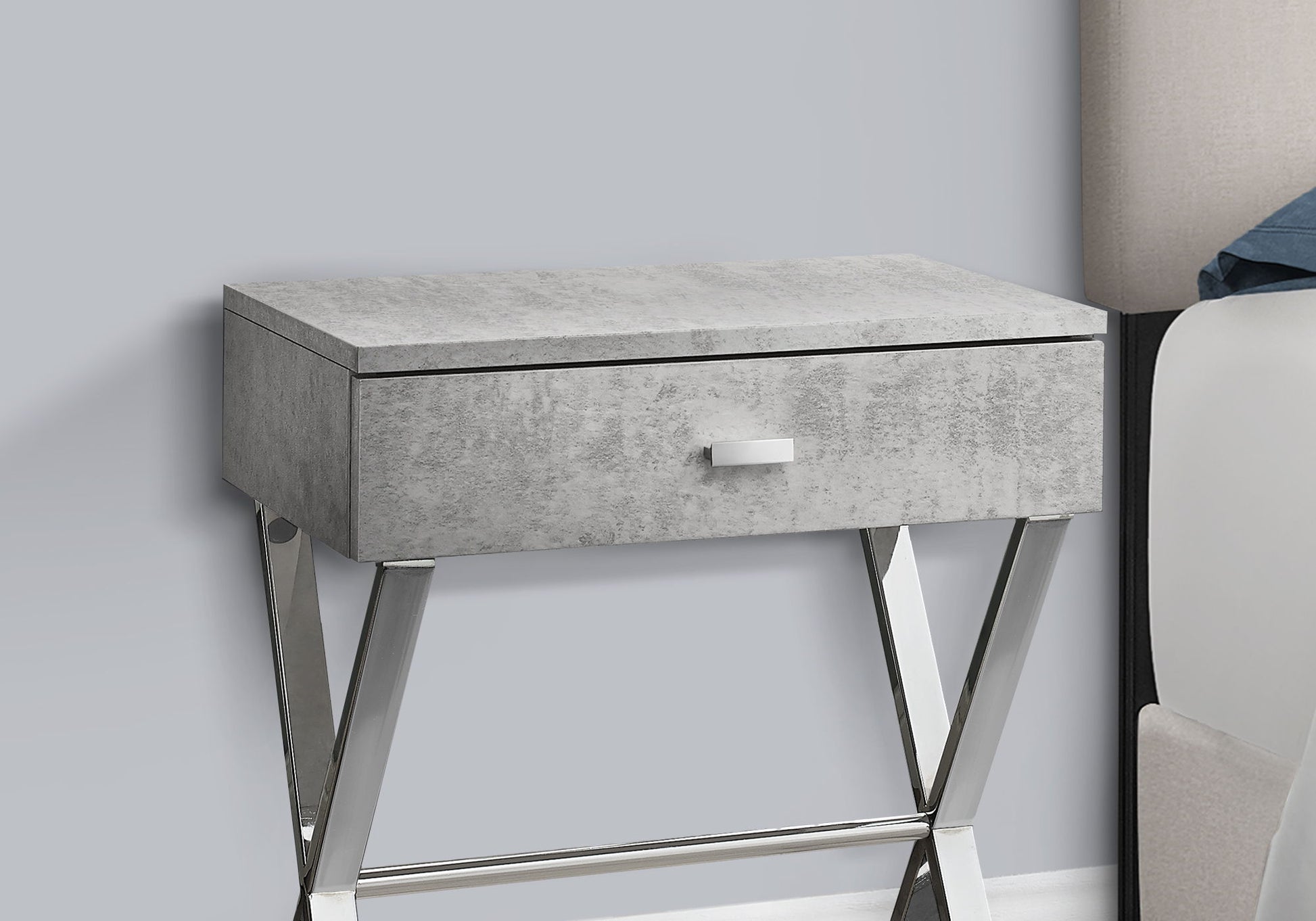 The Accent Side X Table features a gray textured finish, one drawer with a white handle, and angled chrome legs, offering contemporary style and storage. Shown beside a bed against a light gray wall.