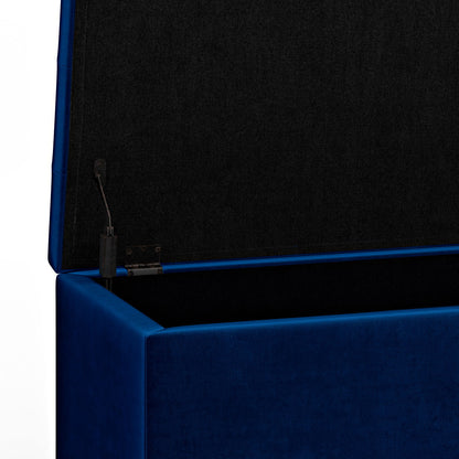 A close-up of the Cosmopolitan - Upholstered Storage Ottoman in blue, open to reveal a black fabric-lined interior and a hinge mechanism on the left side.
