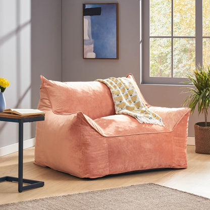 A modern living room featuring the Allea - Bean Bag Chair With Armrests in peach, draped with a patterned blanket, beside a small side table holding yellow flowers, a plant nearby, and sunlight streaming through the window.