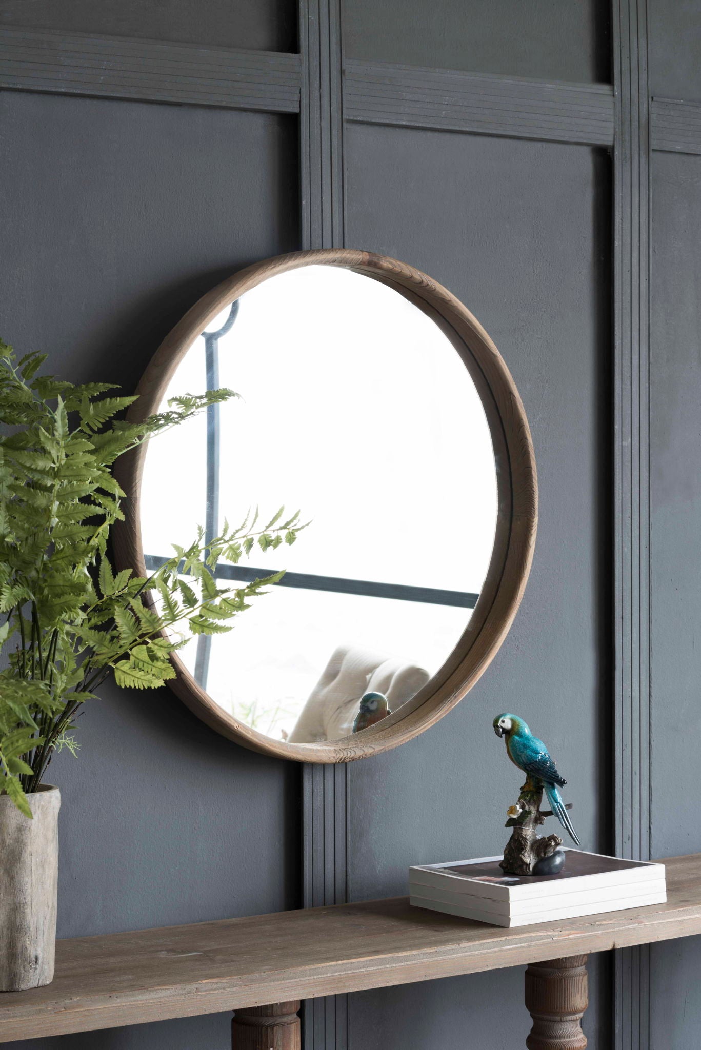 The Round Wood Mirror, Wall Mounted Mirror Home Decor For Bathroom Living Room, hangs on a dark paneled wall above a wooden table with a potted fern, blue parrot figurine, and books. The mirror reflects part of the room.
