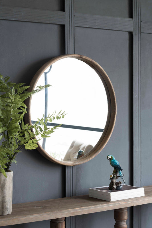 The Round Wood Mirror, Wall Mounted Mirror Home Decor For Bathroom Living Room, hangs on a dark paneled wall above a wooden table with a potted fern, blue parrot figurine, and books. The mirror reflects part of the room.