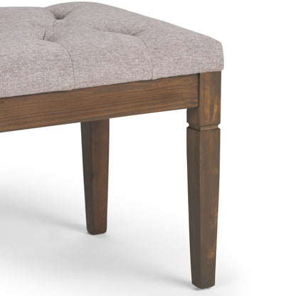 A close-up of the Waverly - Upholstered Tufted Ottoman Bench shows its light gray tufted cushion and dark-stained wooden legs, with only part of the seat and three legs visible.