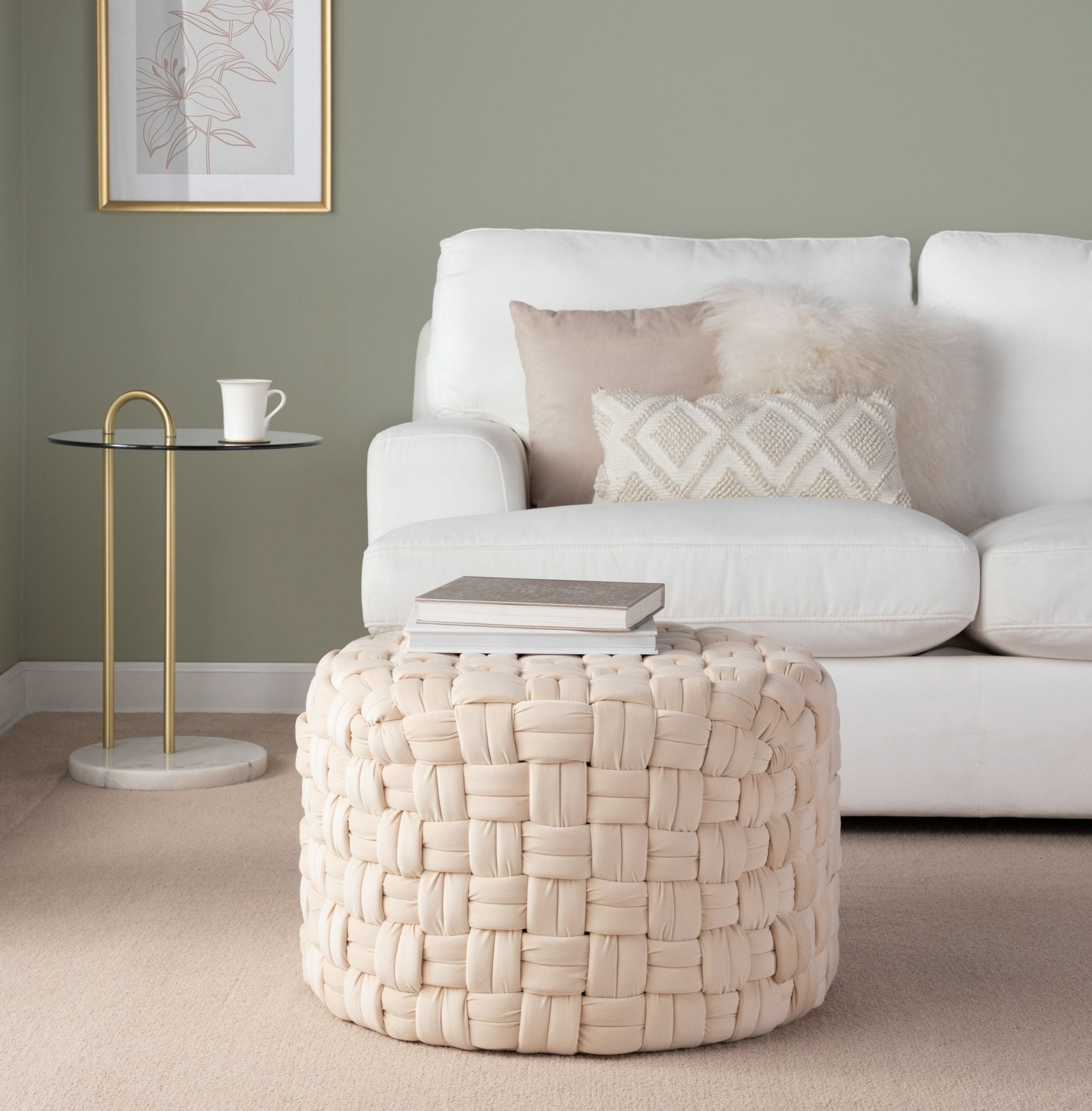 A cozy living room features a white sofa with beige and textured pillows, a Braided - Versatile Round Ottoman, a modern side table with a cup, and framed wall art against a light green wall.