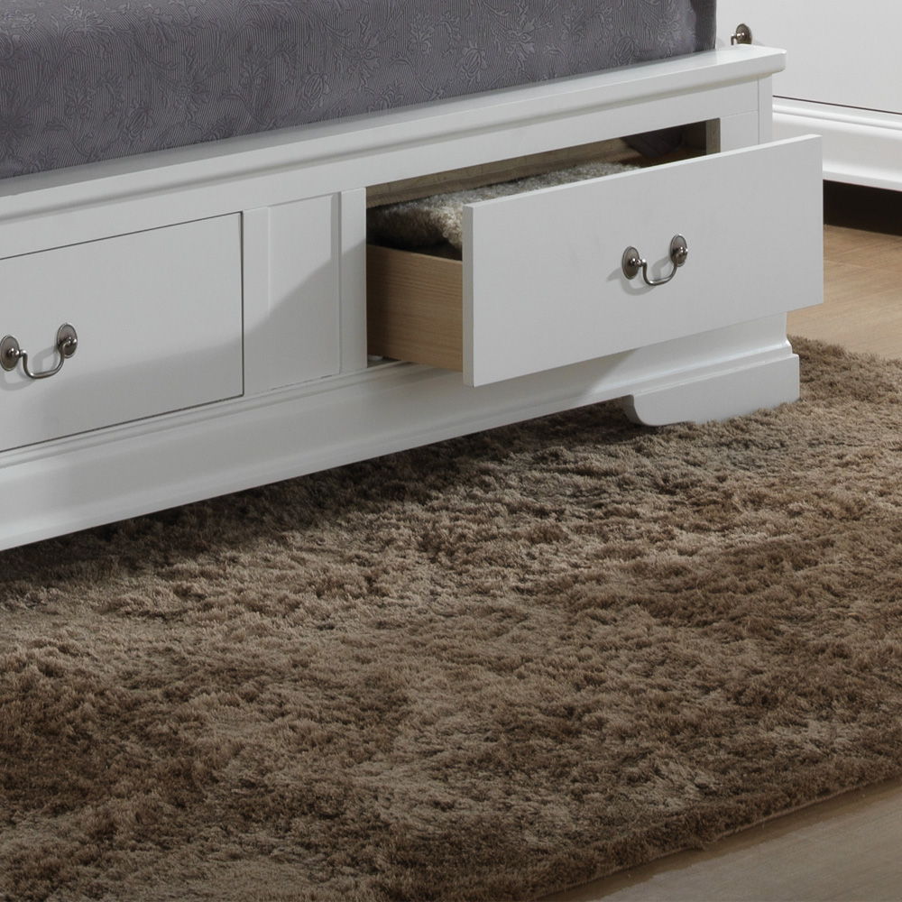 A close-up of the Louis Phillipe - Storage Bed shows its white wooden frame with a storage drawer partially open, revealing a gray rug beneath. The bed features a gray mattress and sits on a fluffy brown carpet.
