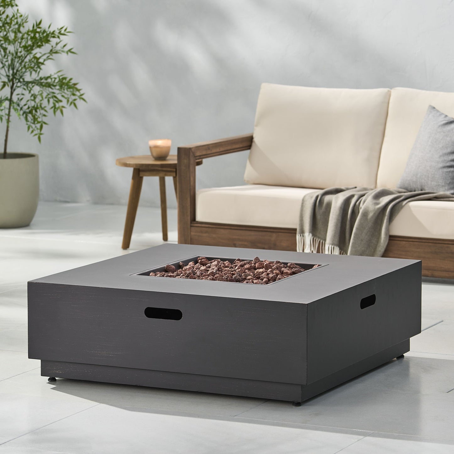 A modern outdoor patio featuring the Modern Square Fire Pit With 50,000 Btu filled with lava rocks, a wooden sofa with cream cushions and a gray throw, a small side table, a potted plant, and a candle.