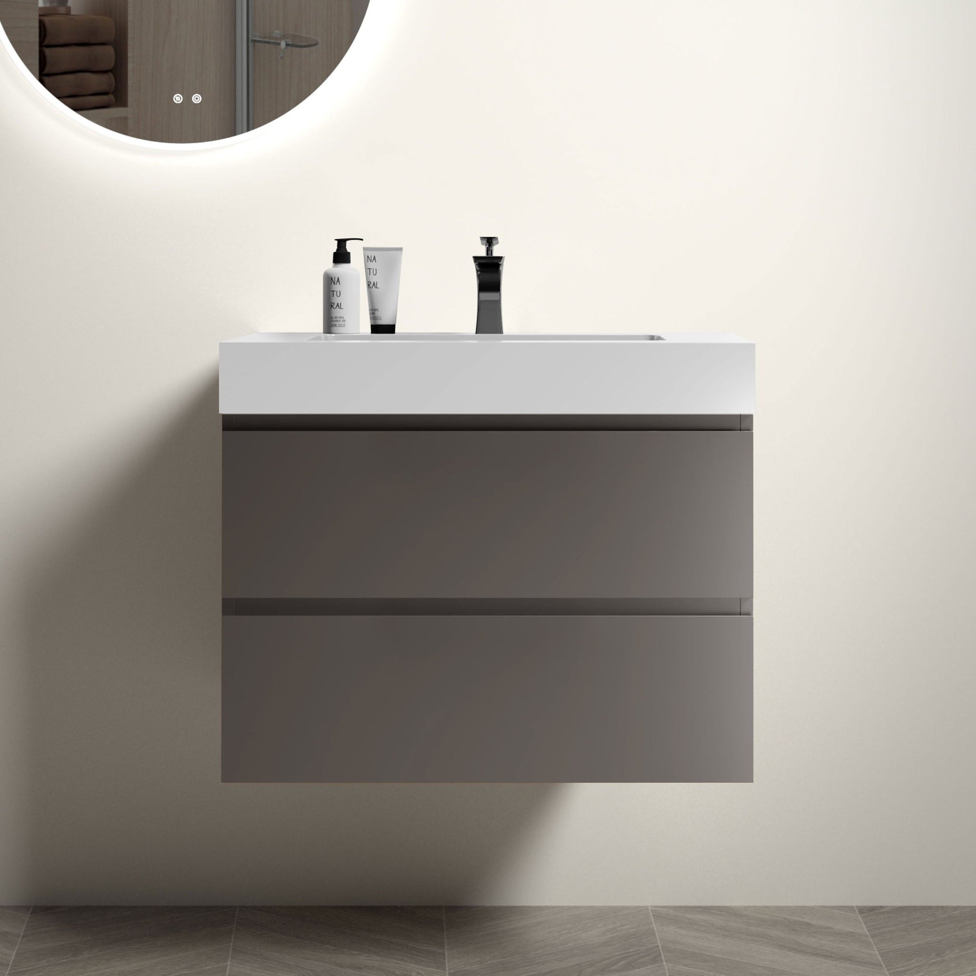 The Alice wall-mounted bathroom vanity features two gray drawers, a white one-piece sink basin (no drain or faucet), and ample storage. Above, a round illuminated mirror reflects towels on a shelf; soap dispensers complete the modern look.