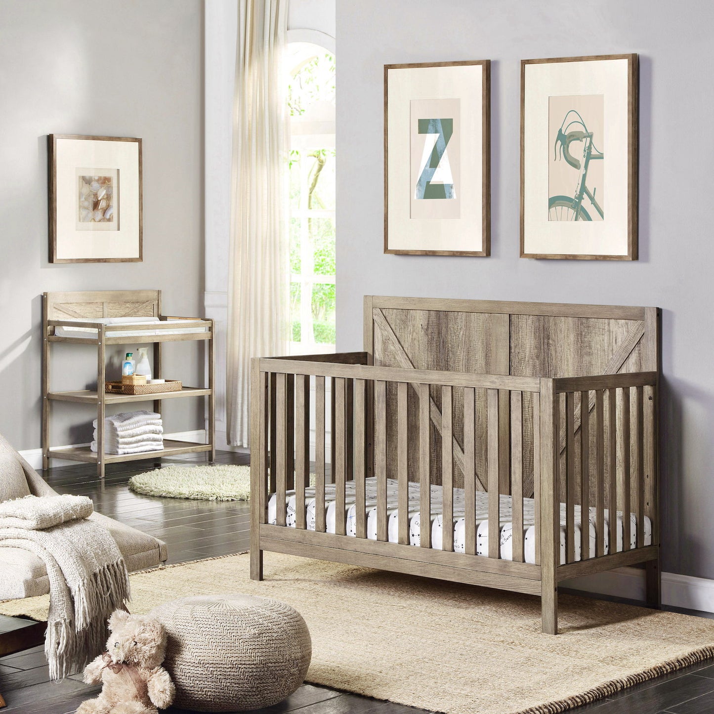 A modern nursery with a wooden crib, soft rug, and framed artwork on light gray walls features the Barnside - Changing Table Vintage. Natural light from a tall window creates a cozy, inviting atmosphere.