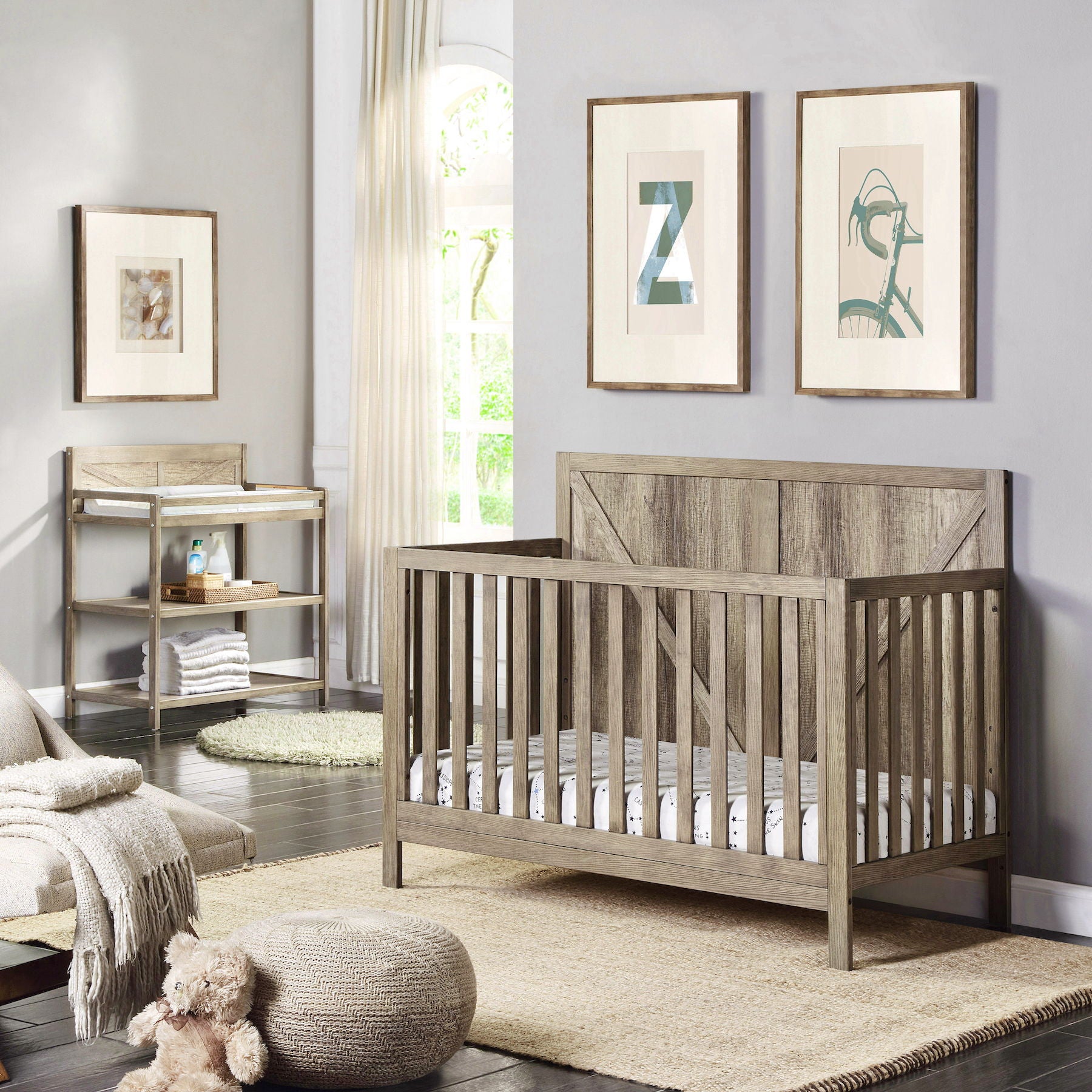 A modern nursery with a wooden crib, soft rug, and framed artwork on light gray walls features the Barnside - Changing Table Vintage. Natural light from a tall window creates a cozy, inviting atmosphere.