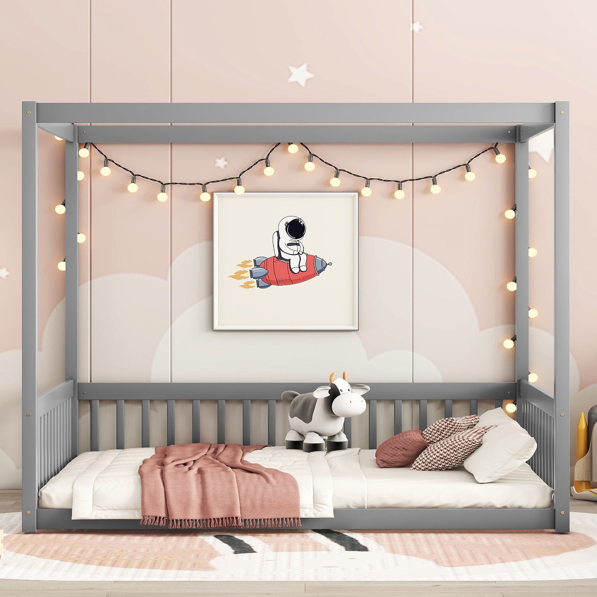 A Canopy Frame Floor Bed With Fence, Guardrails in gray features string lights and astronaut rocket wall art. Cream and pink bedding with a plush cow toy rests on the bed, surrounded by soft, cloud-themed decor.