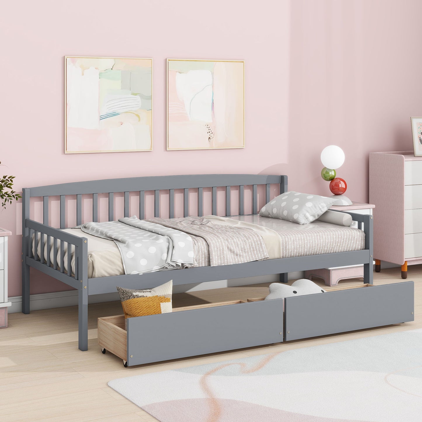 A modern bedroom featuring a Pine Wood Daybed with two storage drawers and a 10-slat bed platform, polka dot comforter, pink walls, abstract artwork, a white nightstand with lamp, and a dresser in the corner.