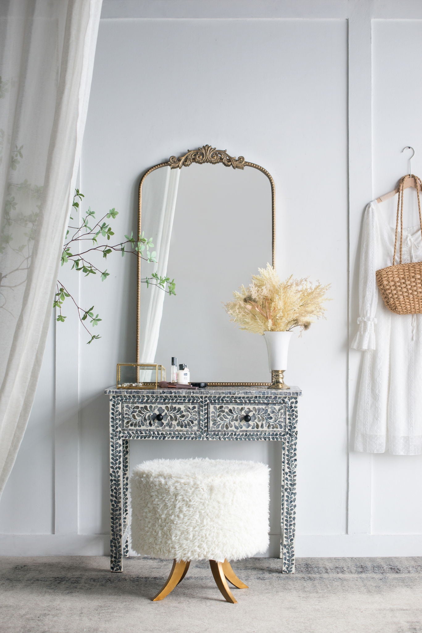 A Baroque-inspired Arch Mirror hangs above an ornate vanity with a fluffy white stool. A vase with dried flowers, beauty products, a hanging white dress, and a wicker purse complete the stylish scene against the white wall.