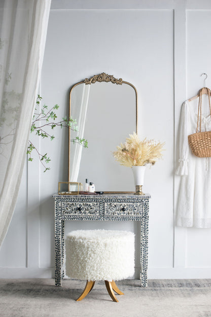 A Baroque-inspired Arch Mirror hangs above an ornate vanity with a fluffy white stool. A vase with dried flowers, beauty products, a hanging white dress, and a wicker purse complete the stylish scene against the white wall.