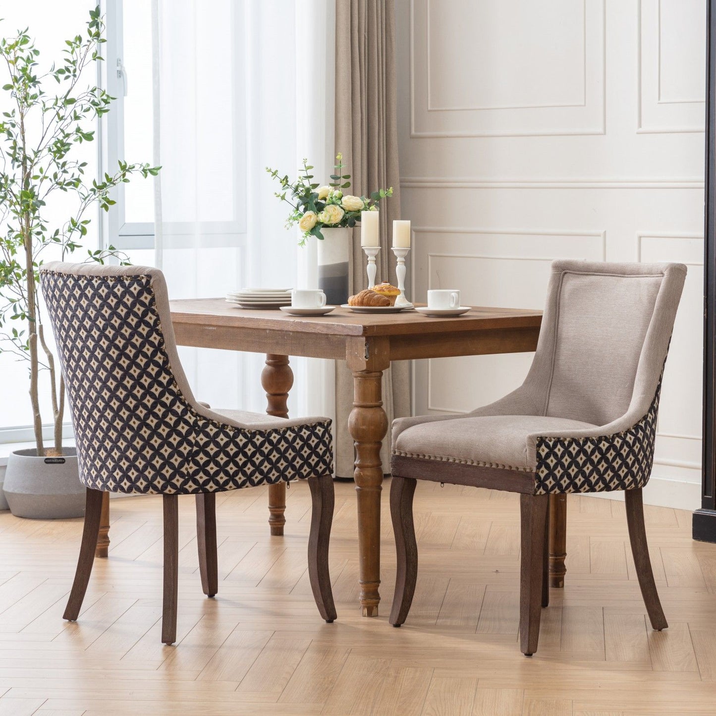 A pair of Ultra Side Dining Chairs with thickened fabric and solid wood legs sits by a windowed table set for two, featuring cups, plates, flowers, candles, and croissants in a bright modern room with plants and curtains.