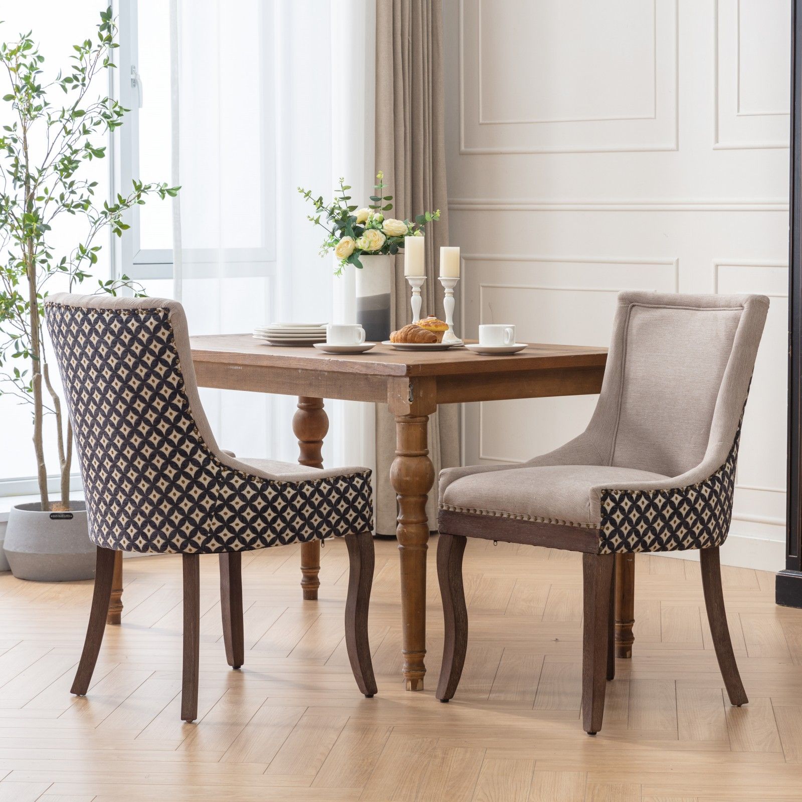 A pair of Ultra Side Dining Chairs with thickened fabric and solid wood legs sits by a windowed table set for two, featuring cups, plates, flowers, candles, and croissants in a bright modern room with plants and curtains.