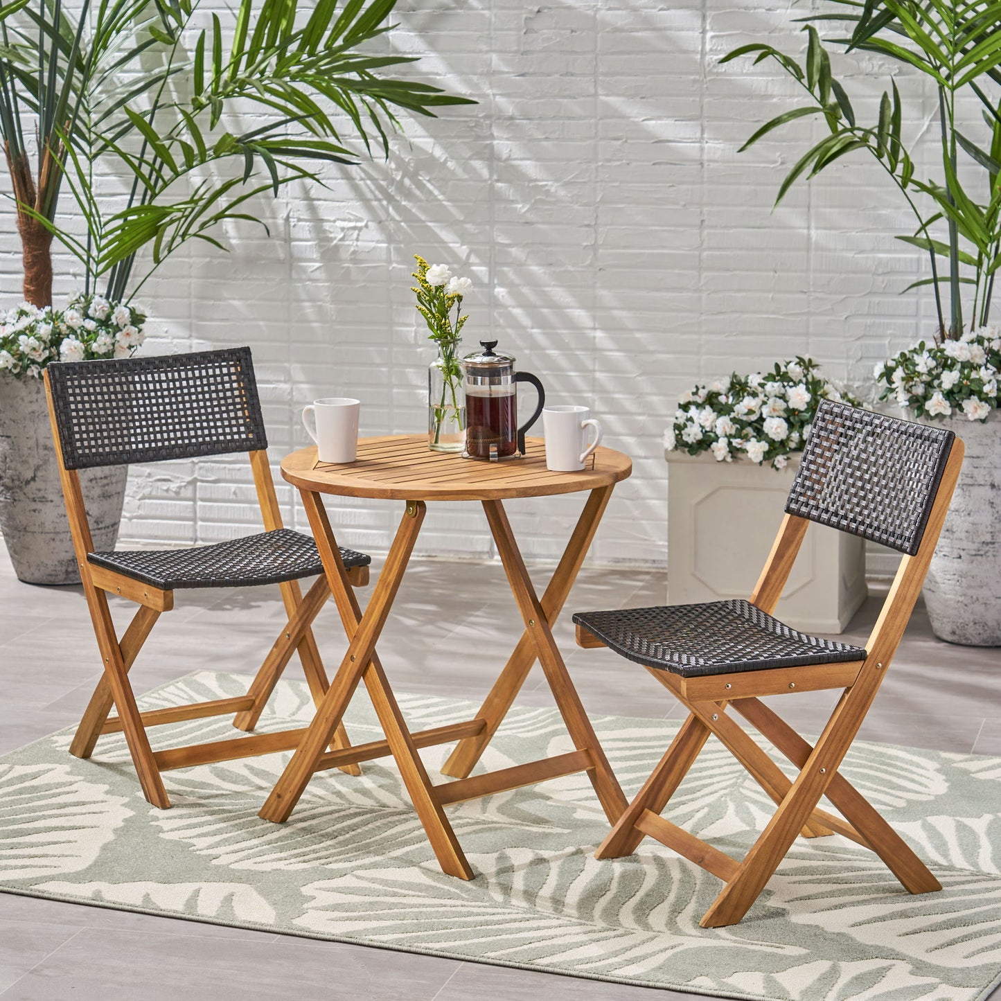 The Evergreen Outdoor Dining Set features a round table and two chairs with woven seats, topped with a French press, two mugs, and flowers in a vase, all set on a patterned rug among potted plants.