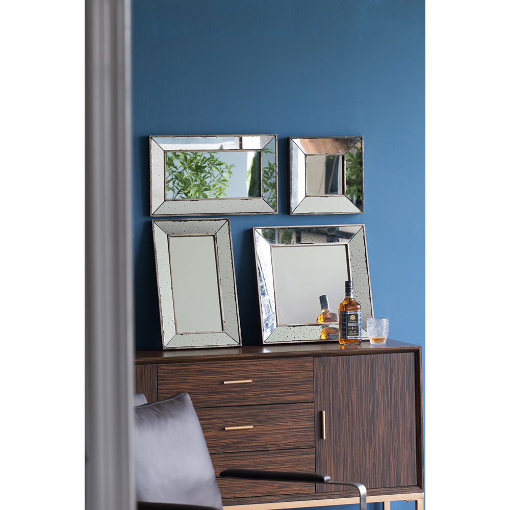 A wooden cabinet against a blue wall displays three Distressed Accent Mirrors, Traditional Style Framed Wall Mirrors for the living room, along with a bottle of whiskey and a glass. A gray cushioned chair is partially visible in the foreground.