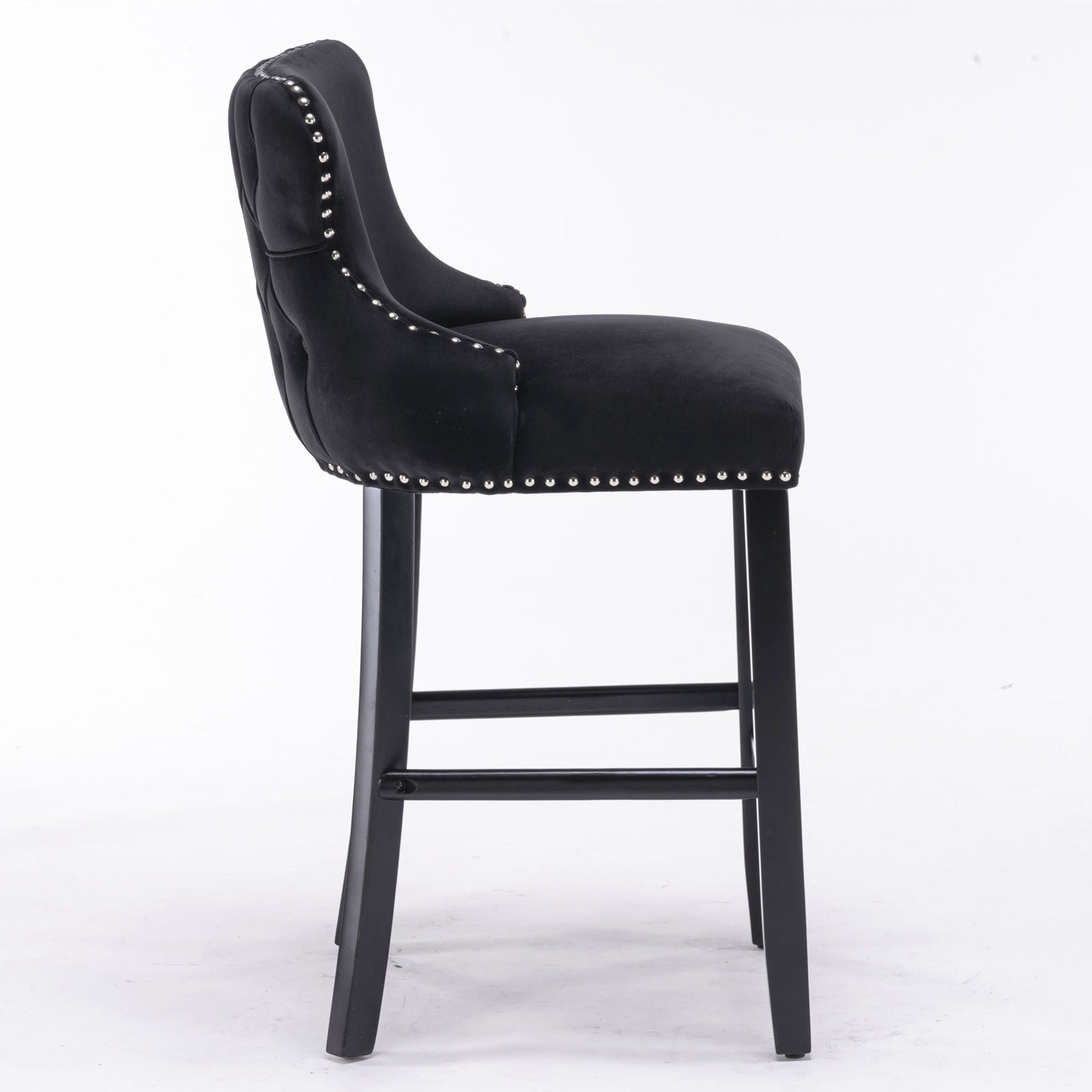 A set of 2 contemporary velvet upholstered wing-back barstools with button tufted decoration, wooden legs, chrome nailhead trim, and footrests is shown from the side against a white background.