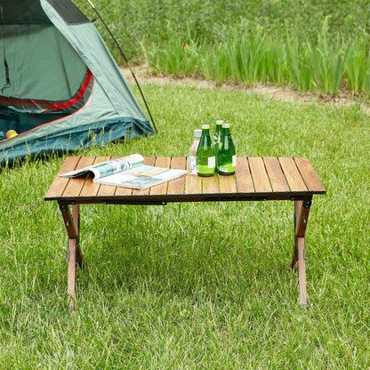 A Folding Outdoor Table, Lightweight Aluminum Roll-Up Rectangular Table in brown holds bottled drinks and an open magazine on grass, with a green tent and tall grass in the background, suggesting a camping scene.