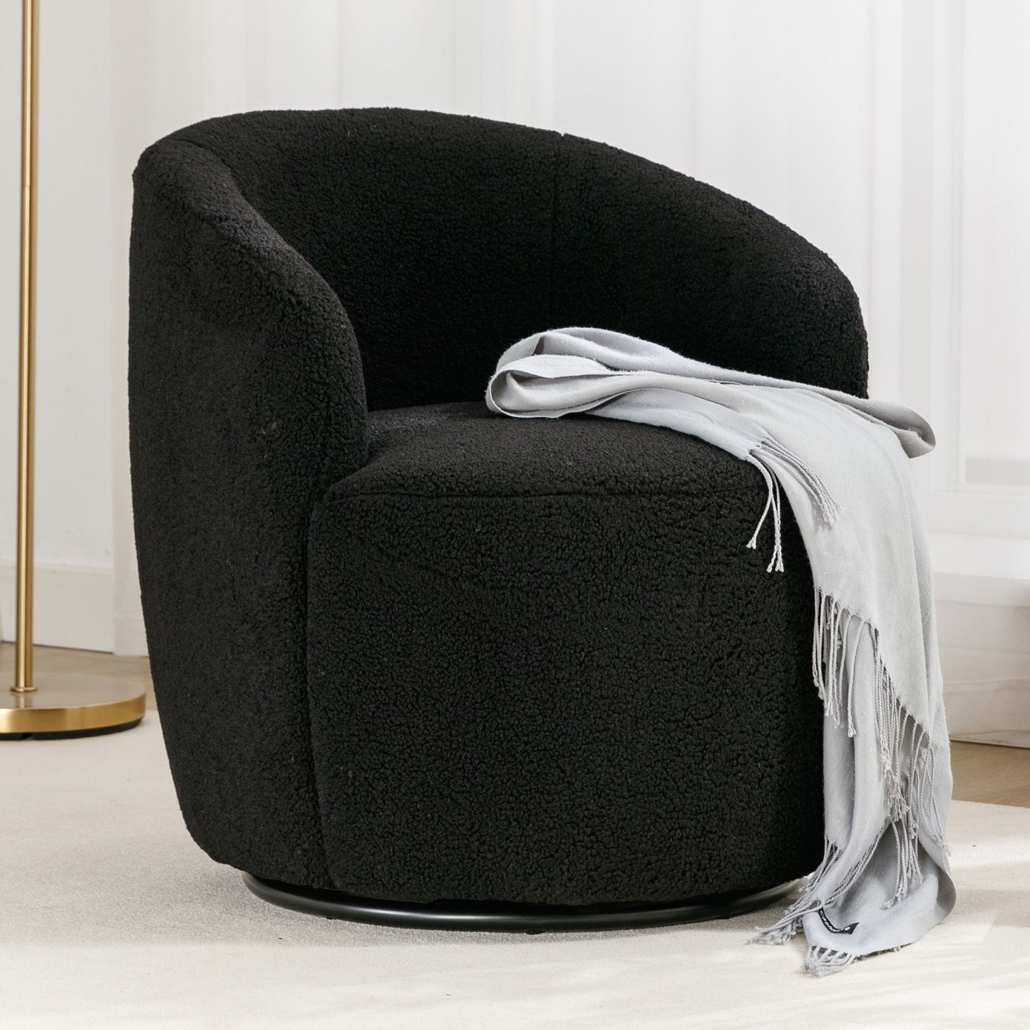 The Teddy Fabric Swivel Accent Armchair Barrel Chair with powder coated metal ring, shown in black textured fabric, is paired with a light gray fringed throw. Placed on a light rug near white curtains and a gold floor lamp.