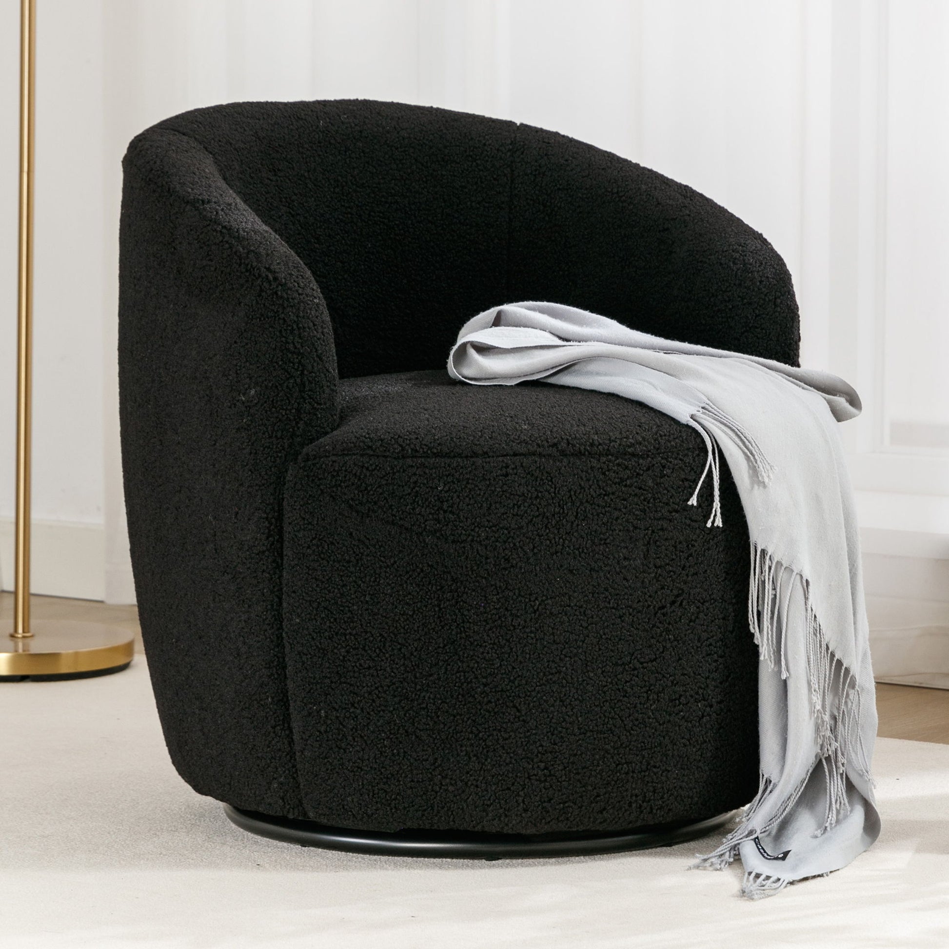 The Teddy Fabric Swivel Accent Armchair Barrel Chair with powder coated metal ring, shown in black textured fabric, is paired with a light gray fringed throw. Placed on a light rug near white curtains and a gold floor lamp.