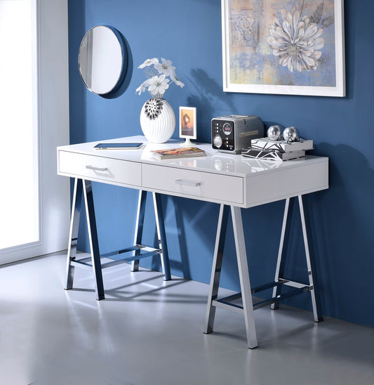 The Coleen High Gloss Writing Desk With USB, finished in white with chrome legs, stands against a blue wall and is styled with white flowers, books, a vintage radio, metallic accents, and topped by a round mirror and floral artwork.