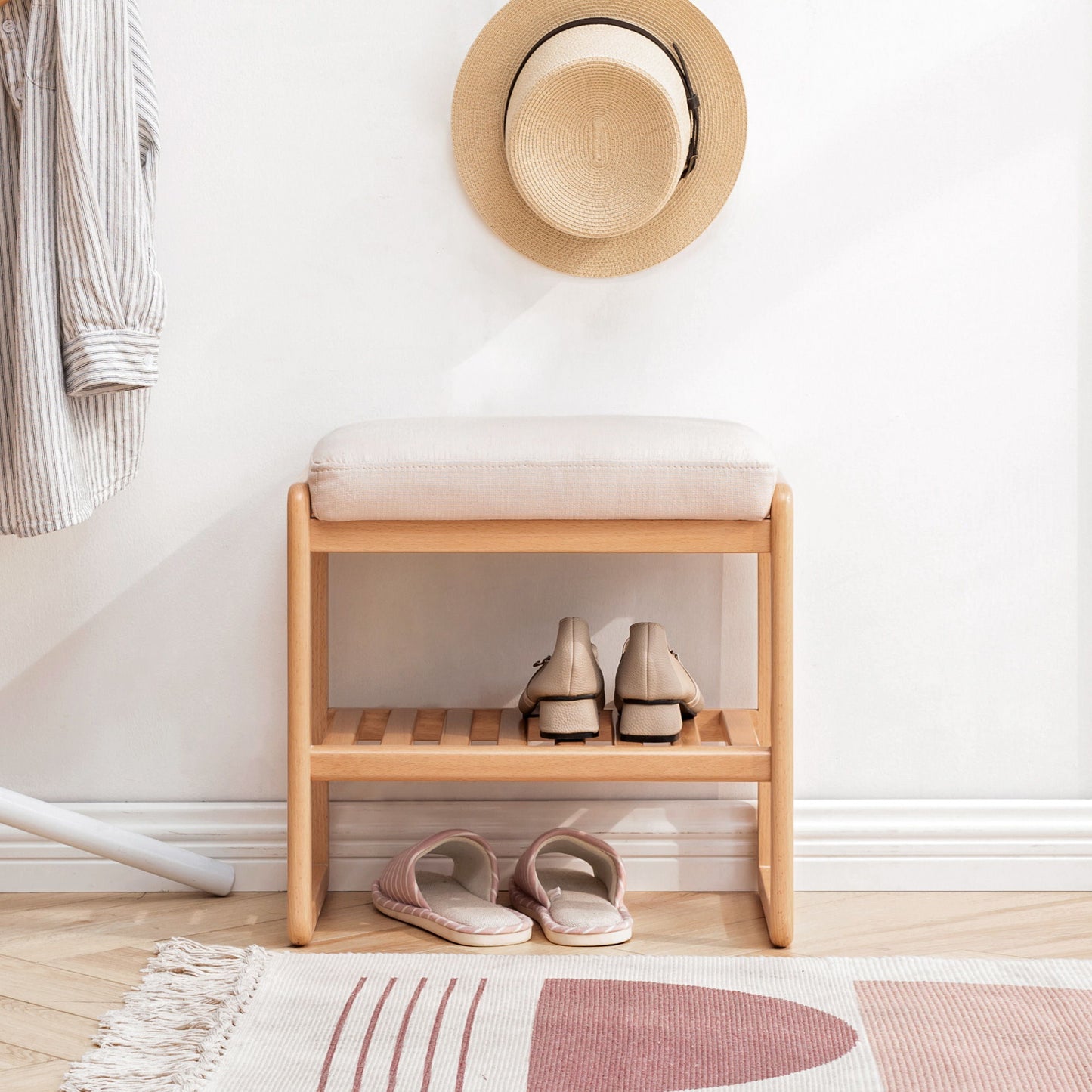 A cozy entryway features the Shoe Bench, Beech Wood Storage Rack Organizer With High Rebound Sponge Cushion - Natural, with shoes on the bottom shelf, slippers nearby, and a striped shirt and straw hat hanging on a white wall.