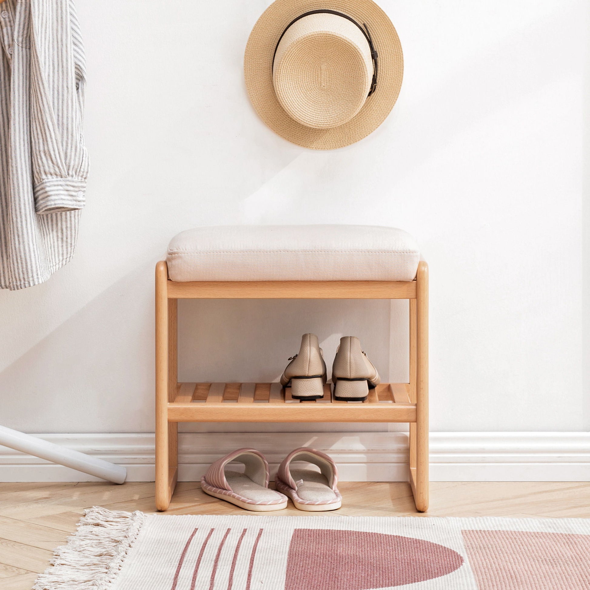 A cozy entryway features the Shoe Bench, Beech Wood Storage Rack Organizer With High Rebound Sponge Cushion - Natural, with shoes on the bottom shelf, slippers nearby, and a striped shirt and straw hat hanging on a white wall.