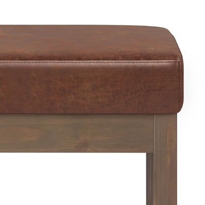 Close-up of the Milltown - Upholstered Ottoman Bench featuring brown faux leather upholstery, wooden frame and legs, and detailed texture, shown against a white background.